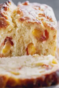 A slice of buttermilk peach bread displayed on a cutting board with fresh peach slices and mint, highlighting its golden brown texture.