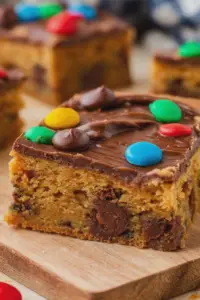 Close-up of a peanut butter cookie bar with chocolate topping and M&M's on a wooden table.