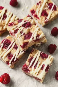 Close-up of raspberry and white chocolate squares on a wooden board with fresh raspberries and white chocolate curls.