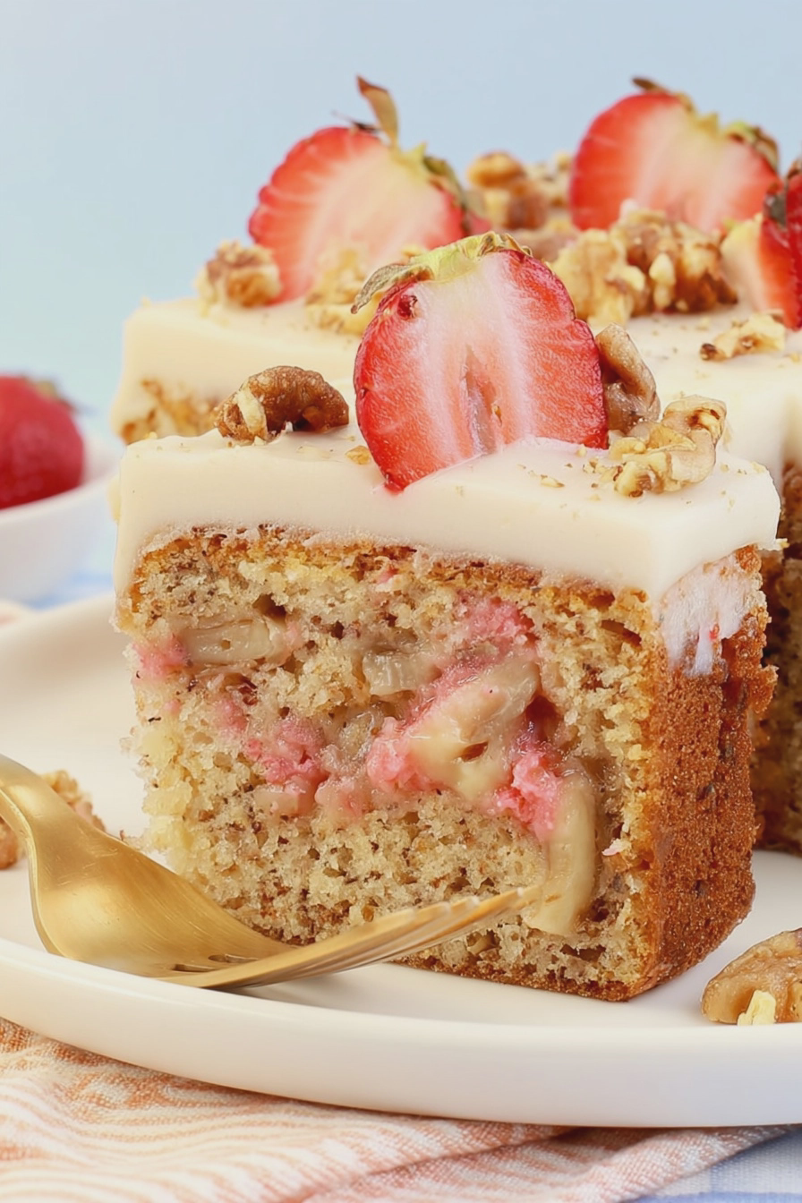 Delicious strawberry banana cake with cream cheese frosting decorated with fresh strawberries and chopped walnuts.