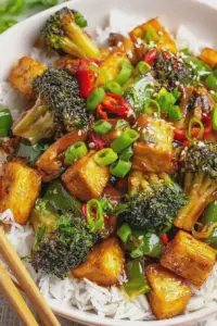 A colorful bowl of tofu stir fry with bell peppers, broccoli, and sesame seeds served over rice, ready to eat.